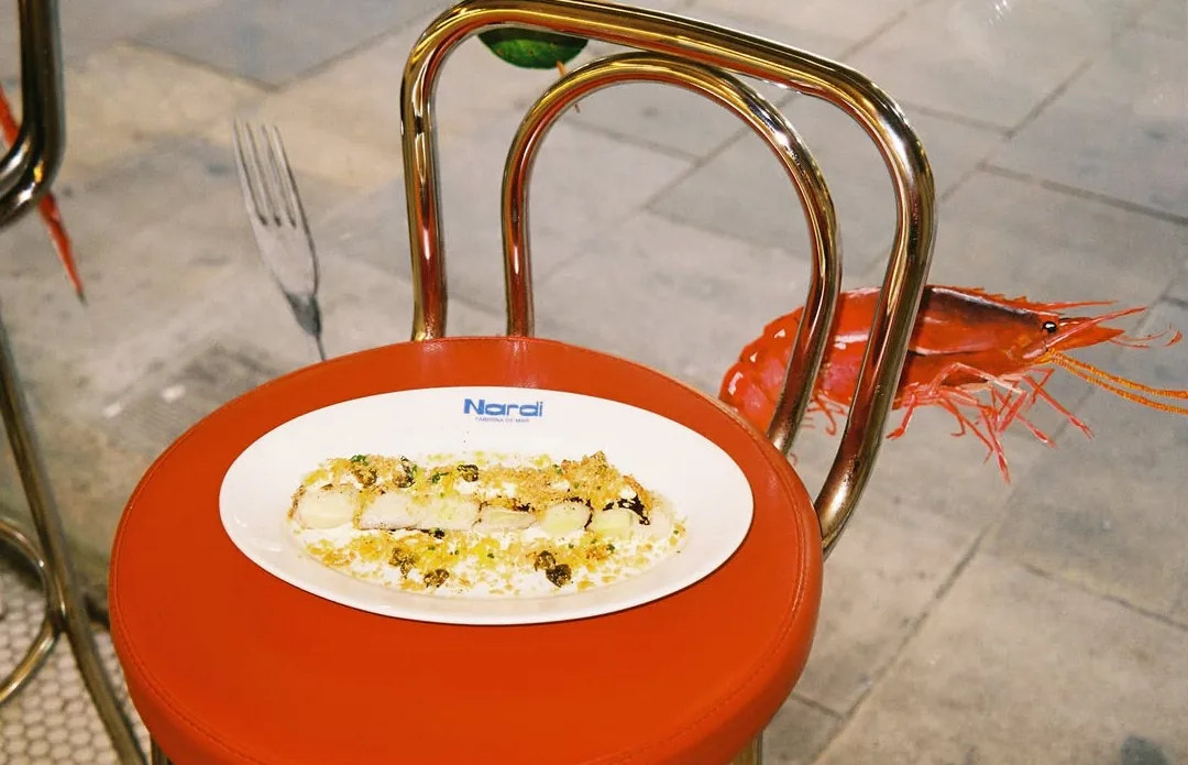Un plato con el logo de Nardi sobre un taburete rojo: pescado con patata, alcaparras y migas