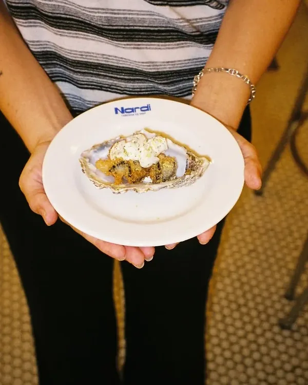 Unas manos sostienen una ostra con su guarnición, en un plato con el logo de Nardi